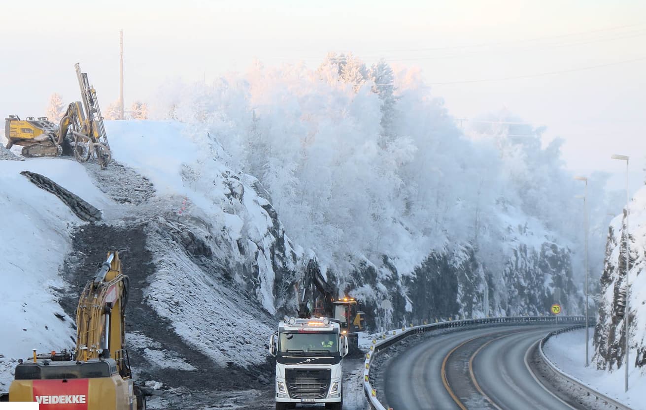Veidekke excavators and drilling equipment working on a snow-covered rocky hillside alongside the E6 motorway in Norway, with a truck navigating the road below in winter conditions.