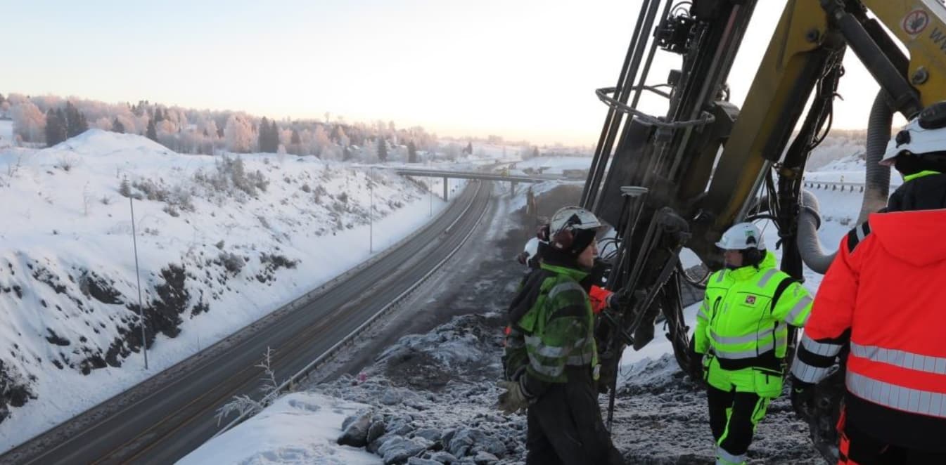 Veidekke construction workers in high-visibility gear operating drilling equipment on a snowy hillside overlooking the E6 Arnkvern-Moelv motorway construction site in Norway.
