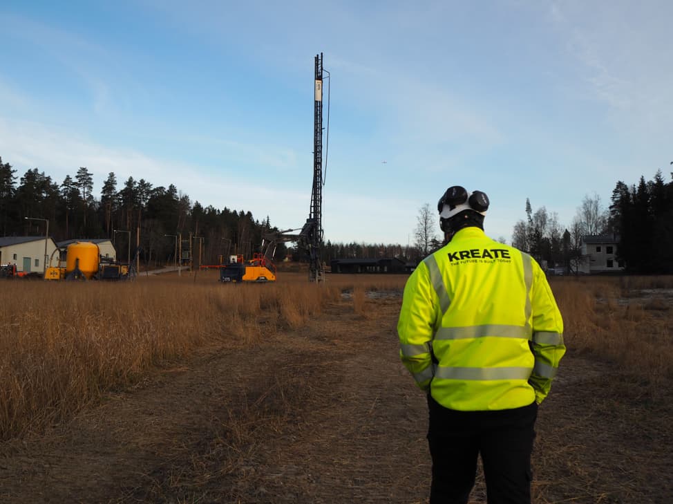 A Kreate Oy worker in a high-visibility jacket overlooking the early-stage Streets of Kallvik construction site in Espoo, with drilling equipment and machinery visible across an open field.