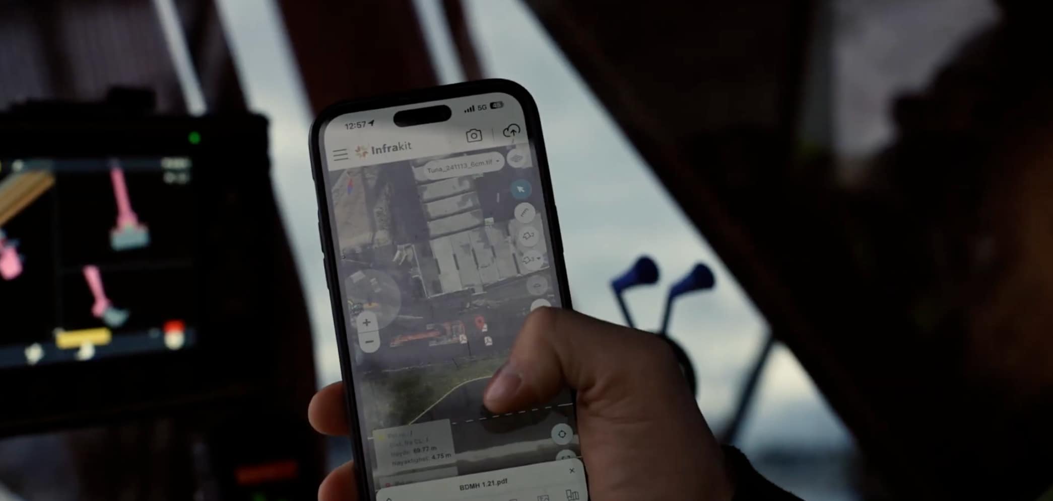 A site worker holding a smartphone running the Infrakit mobile app, showing a map-based view of an active construction site with design data and machine positions.