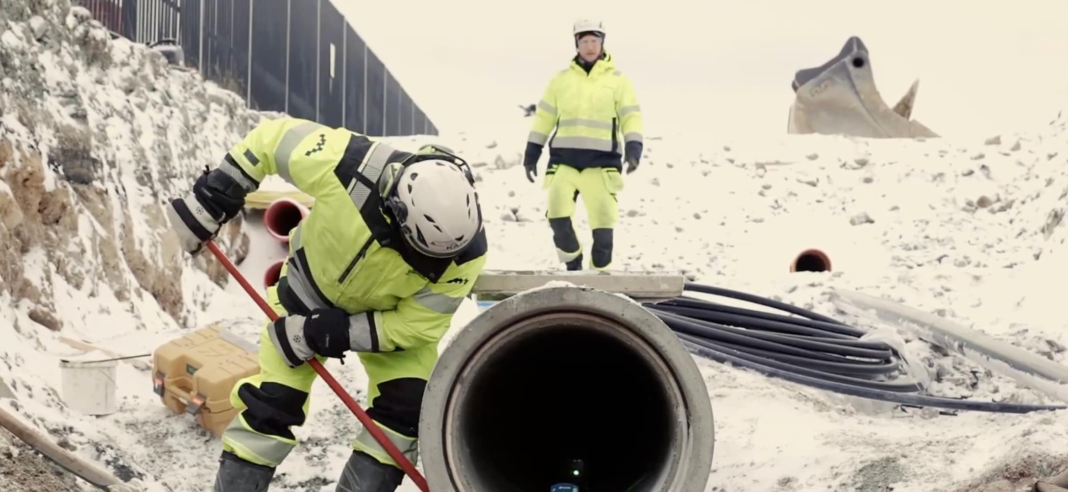 Two construction workers in high-visibility gear handling a large concrete pipe on a snowy civil engineering site, with an excavator bucket visible in the background.