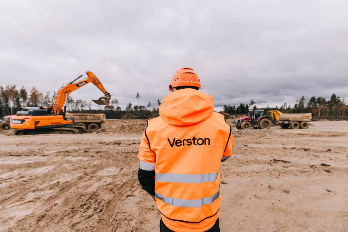 Verston site worker overseeing Rail Baltica earthworks with excavator and trucks in background